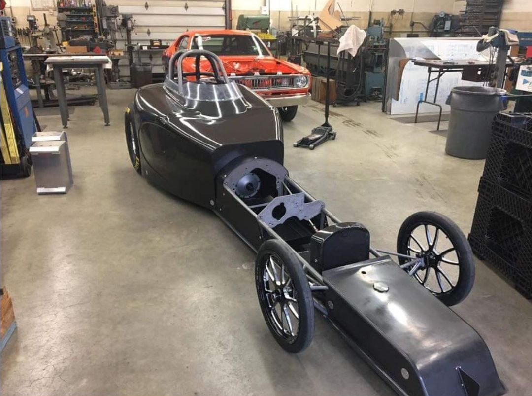 A dragster is sitting in a garage next to a red car
