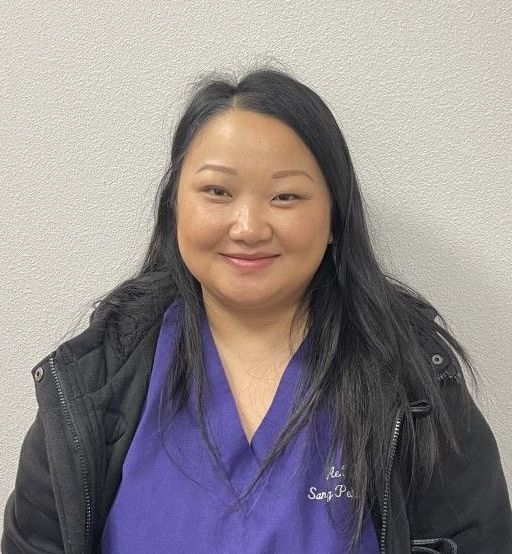A woman wearing a purple scrub top and a black jacket is smiling for the camera.