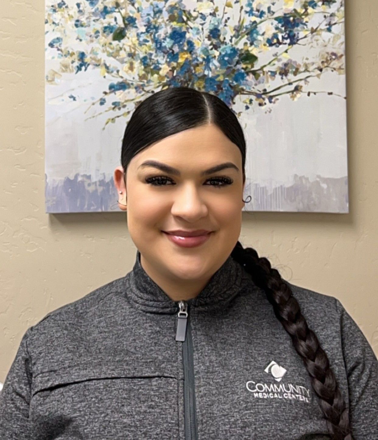 A woman is smiling in front of a painting of flowers on a wall.