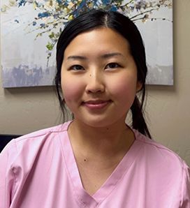 A woman in a pink scrub top is smiling in front of a painting on the wall.