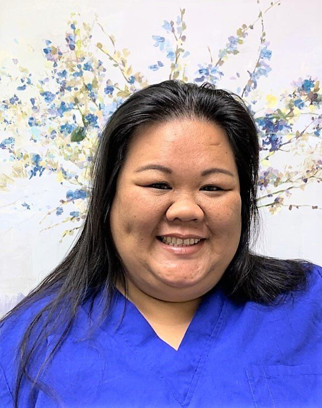 A woman in a blue scrub top is smiling in front of a painting of flowers.