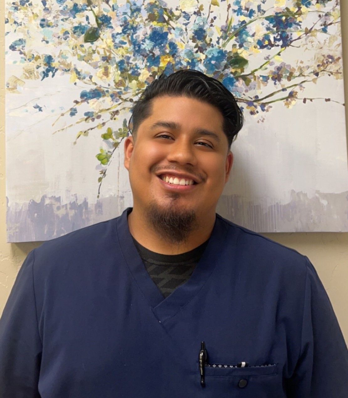 A man in a blue scrub is smiling in front of a painting.