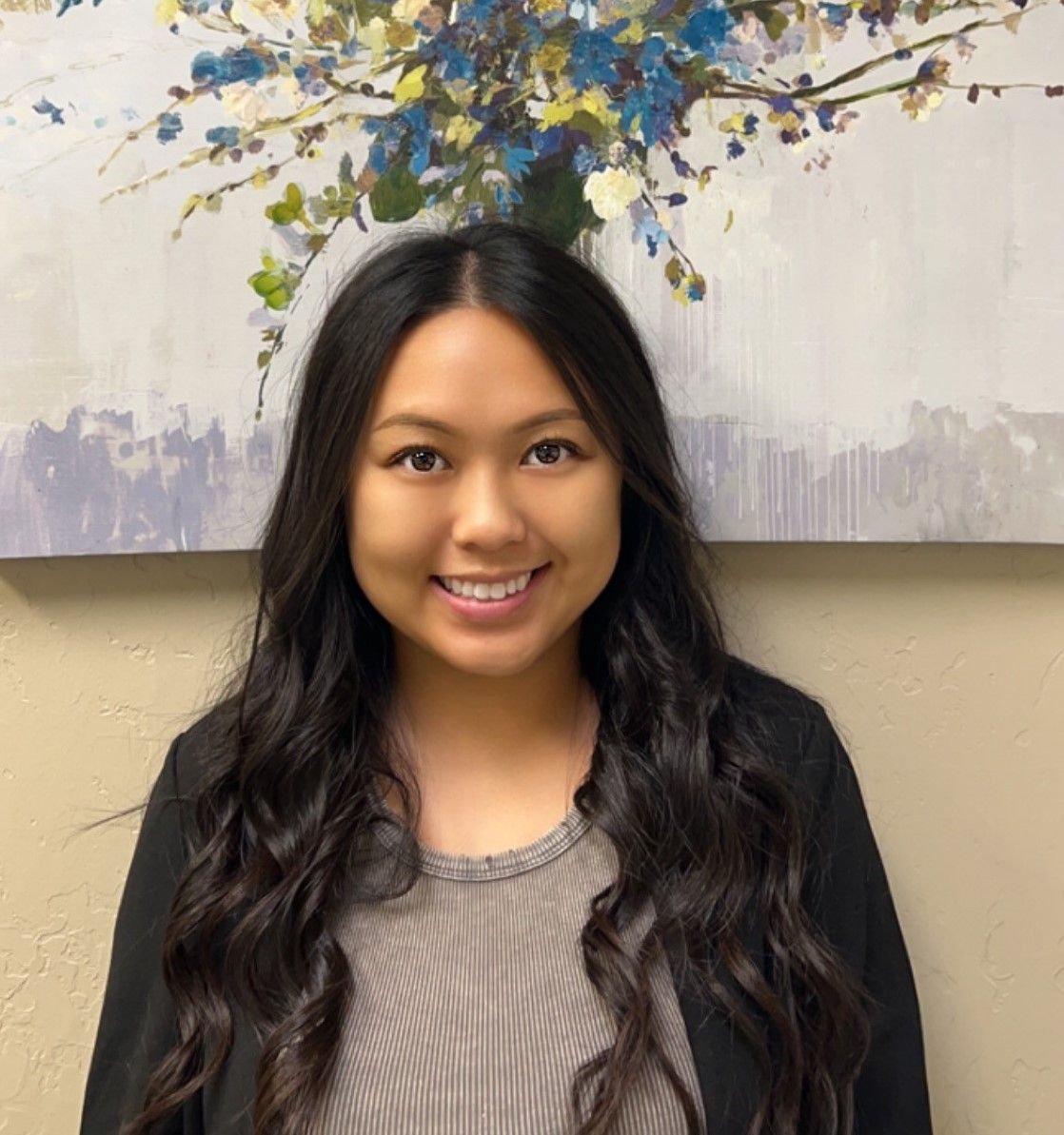A woman is smiling in front of a painting of flowers on a wall.