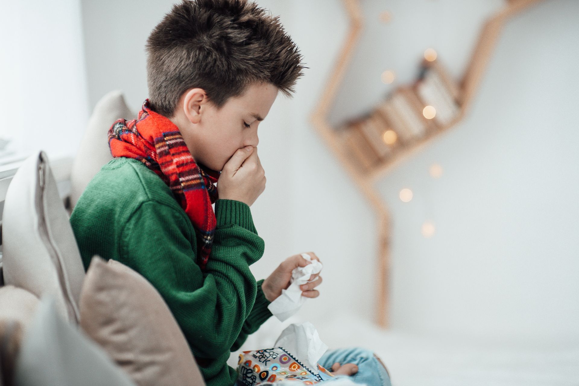 A young boy is sitting on a couch coughing into a napkin.