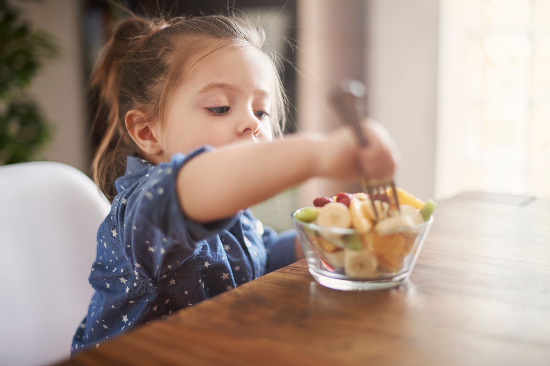 A little girl is sitting at a table eating a fruit salad with a fork.