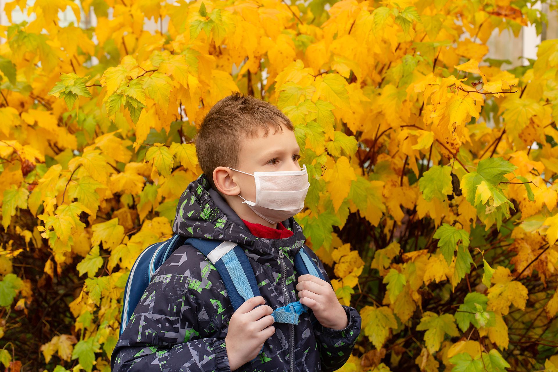 A young boy wearing a mask and a backpack is standing in front of a bush with yellow leaves.