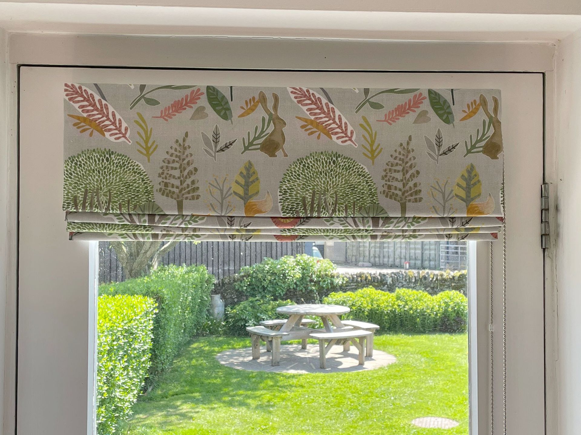A window with a blind on it looking out to a garden with a picnic table.