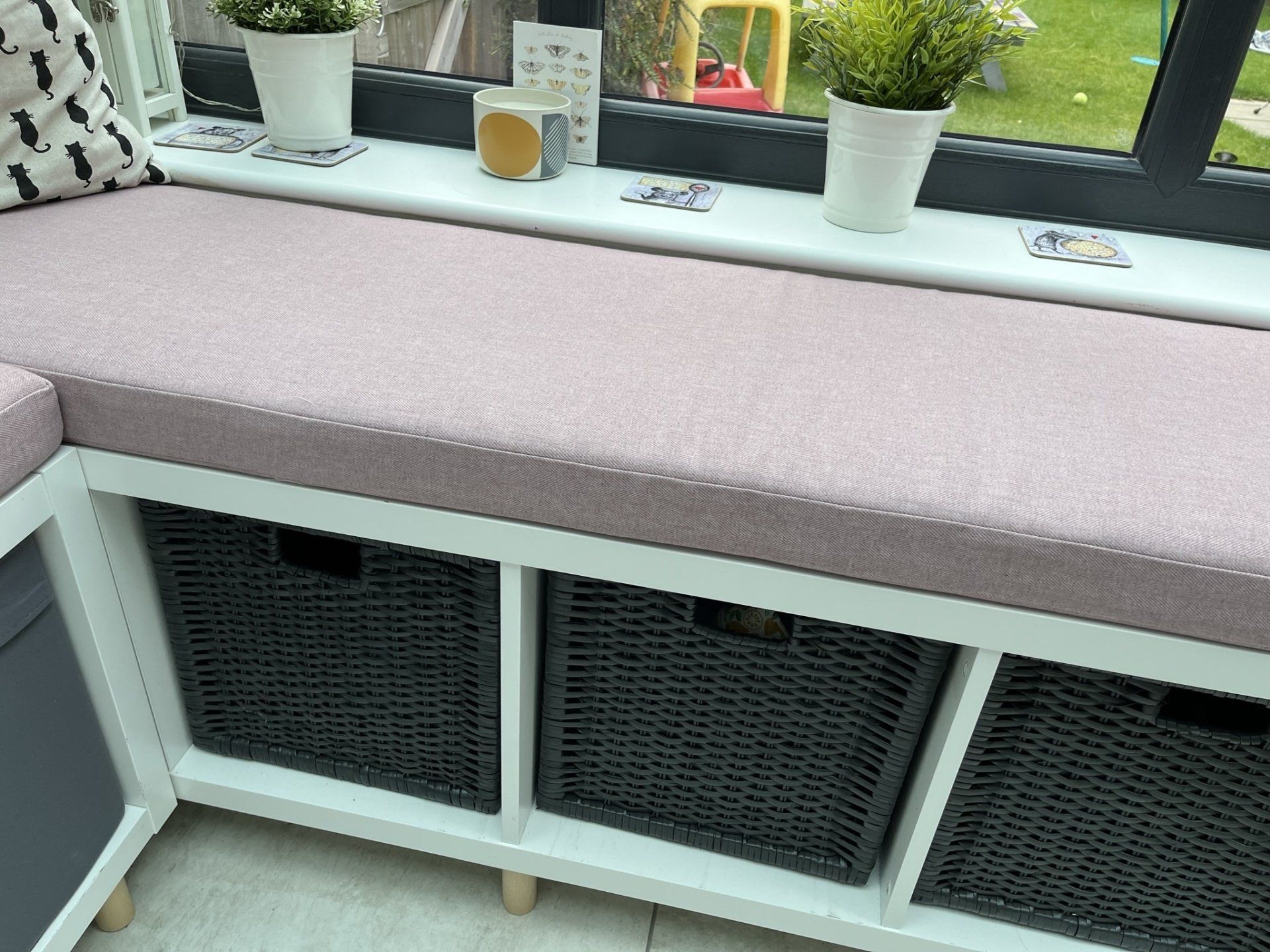 A window seat with a pink cushion and wicker baskets underneath it.
