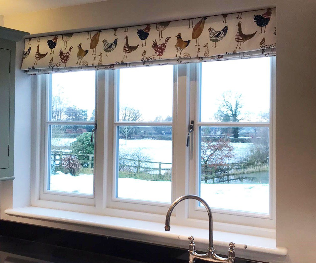 A kitchen with a sink , window , and curtains with chickens on them.
