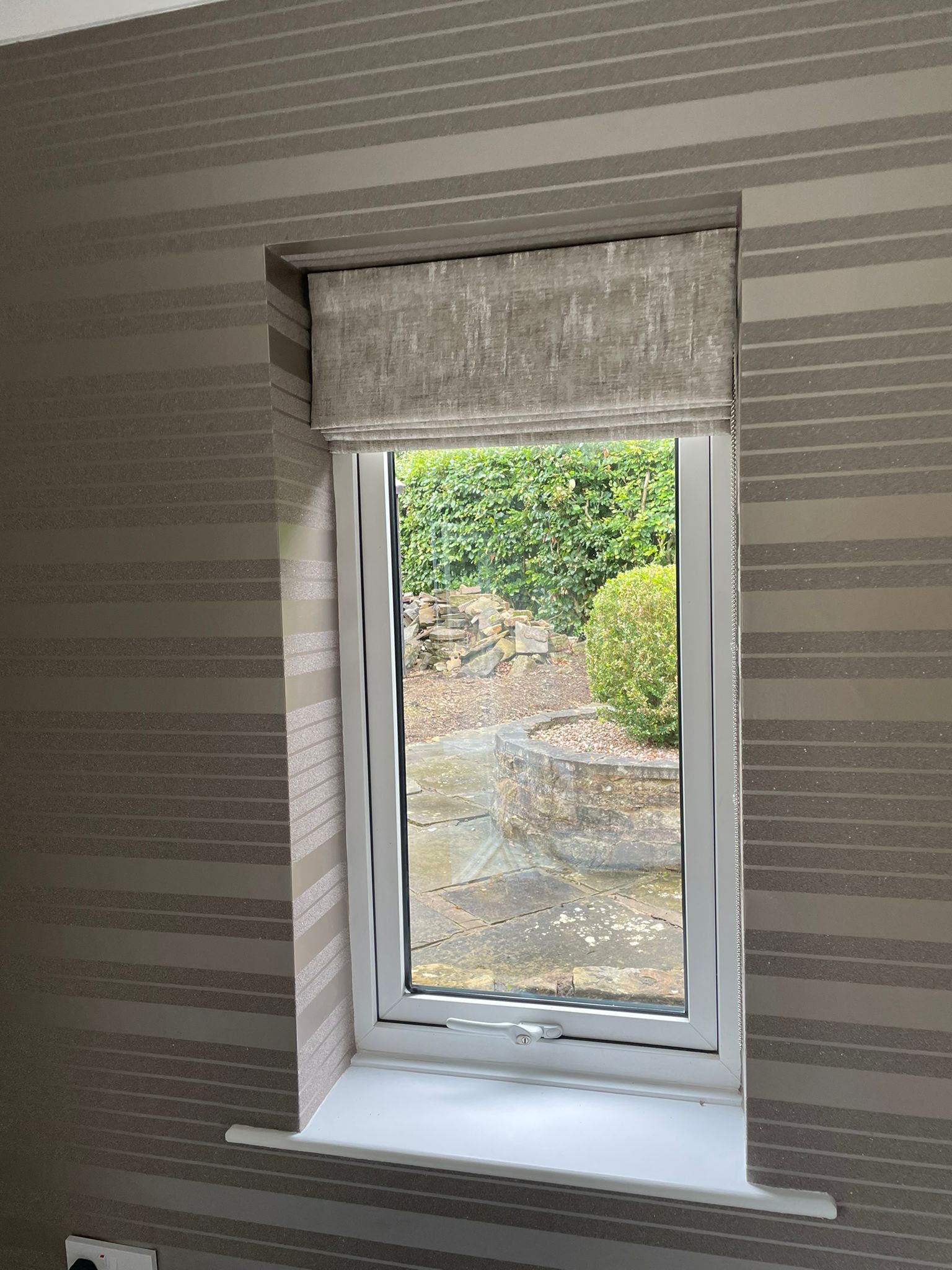A window with a roman blind on it in a room with striped wallpaper.
