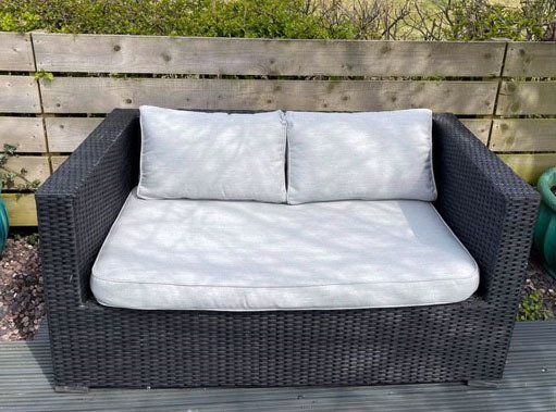 A black wicker couch with white pillows is sitting on a wooden deck.
