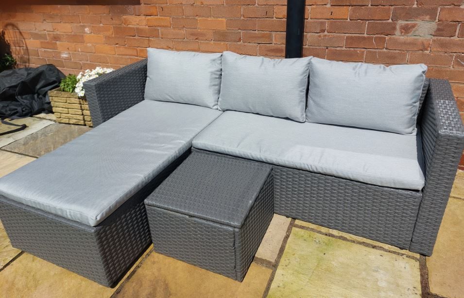 A wicker sectional couch with grey pillows and a table on a patio.