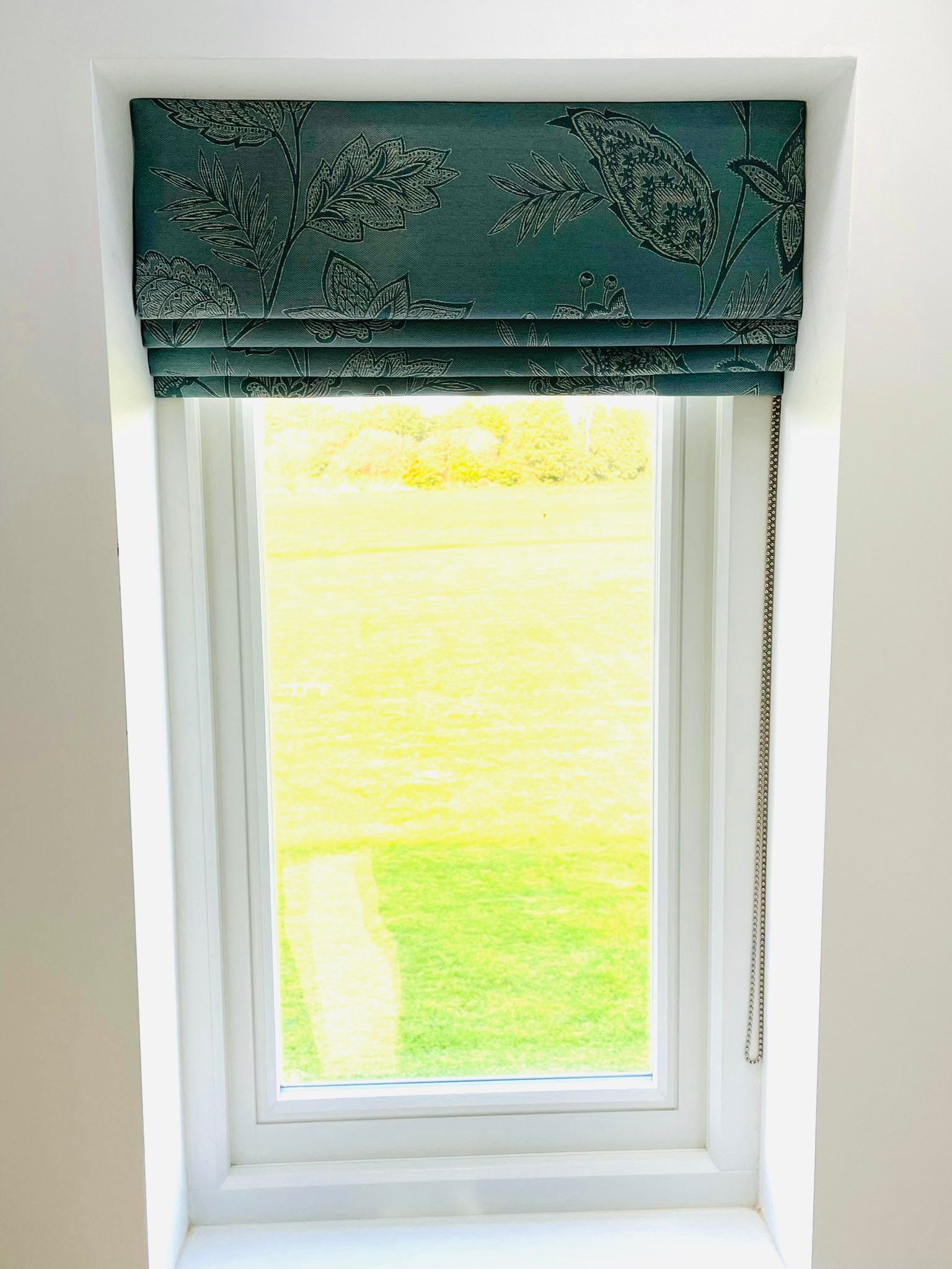 A window with a blue roman shade on it and a view of a field.