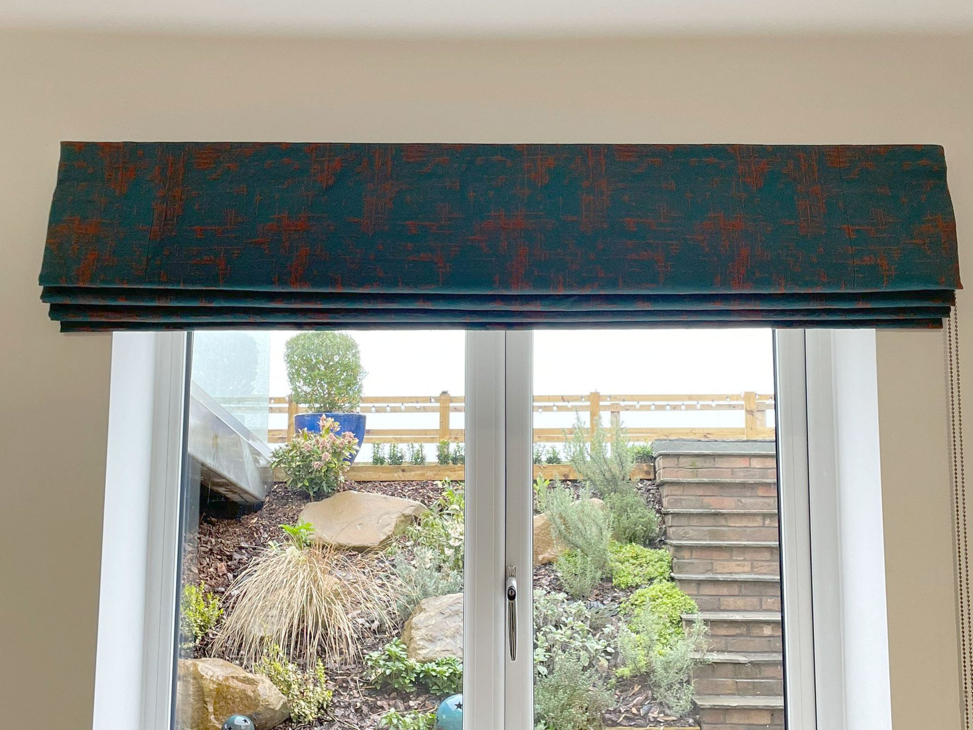 A window with a blue roman blind on it and a view of a garden.