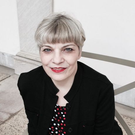 A woman wearing a black jacket and red lipstick smiles for the camera