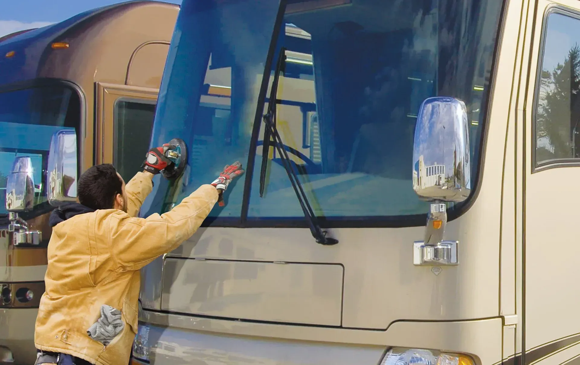 Person cleaning the windshield of a beige RV with a buffer, outdoors on a sunny day.