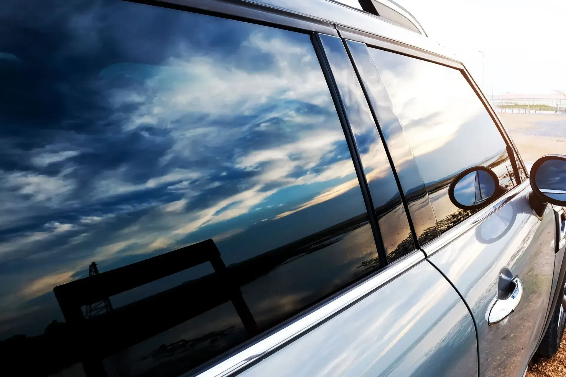 Car window reflecting a cloudy sky, near a beach. Silver car with a side mirror visible.