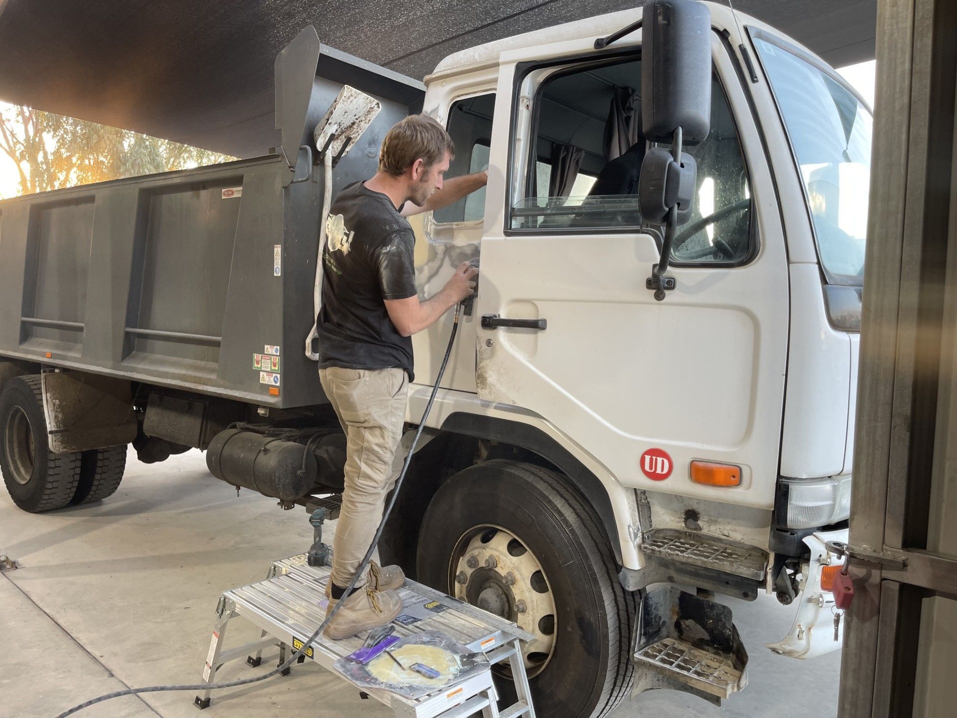 A man works on the door of a white dump truck, standing on a small step stool. The truck is in a garage — Panel Pro Smash Repairs in Wodonga, VIC