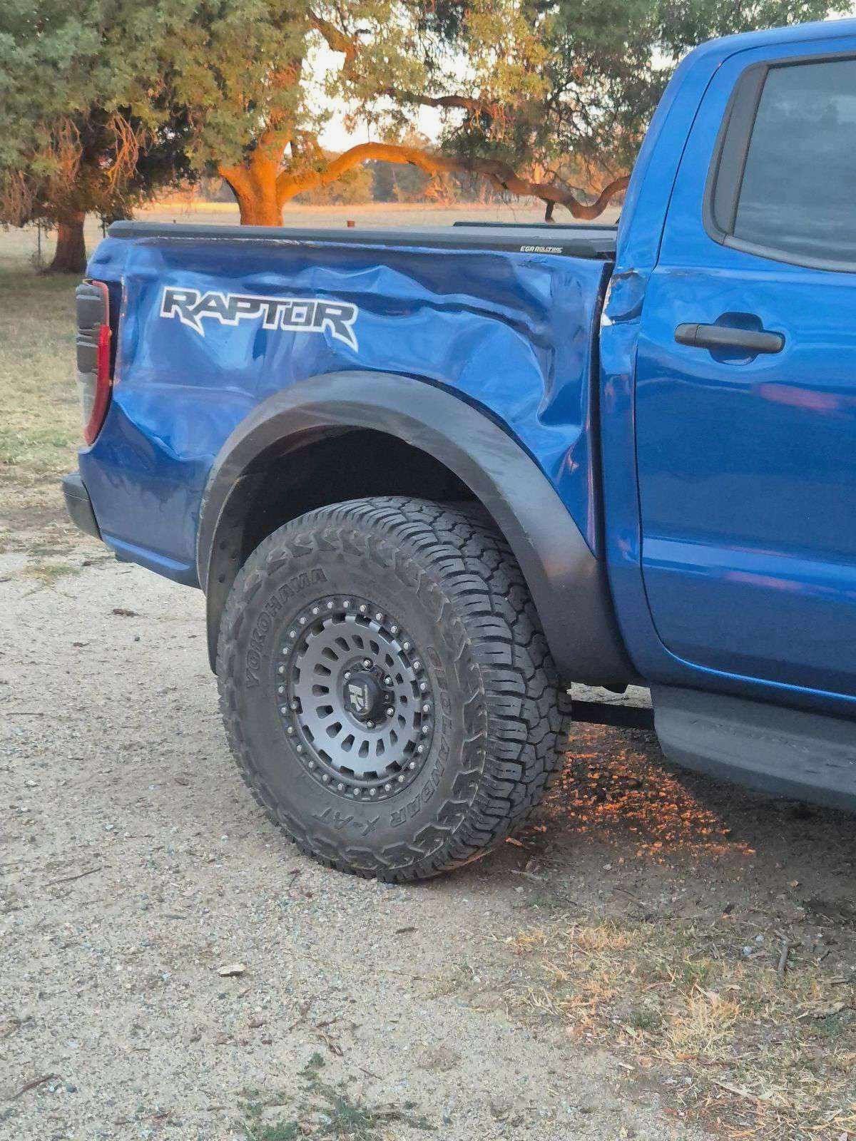 Blue Ford Raptor truck parked on a dirt road with large tires. The truck has black fender flares and 