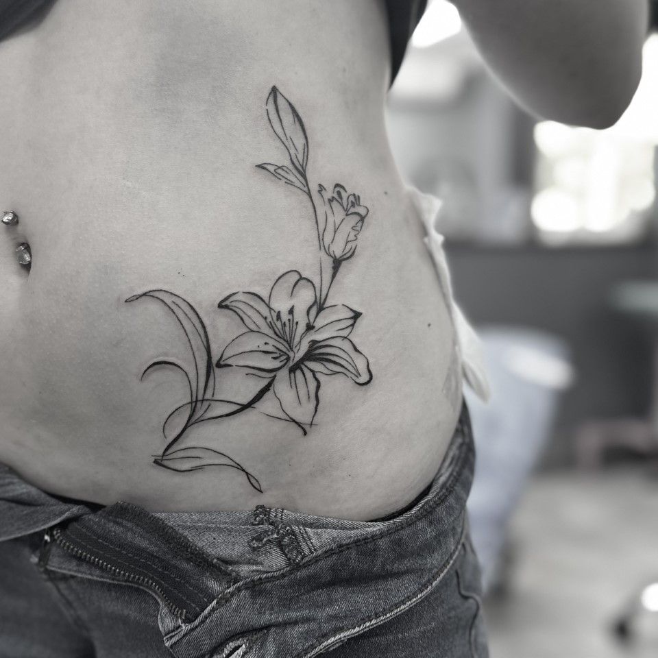 Black ink lily tattoo on abdomen, partially visible in blue jeans. Pierced navel. — Whitsunday Tattooing Airlie Beach in Whitsundays, QLD