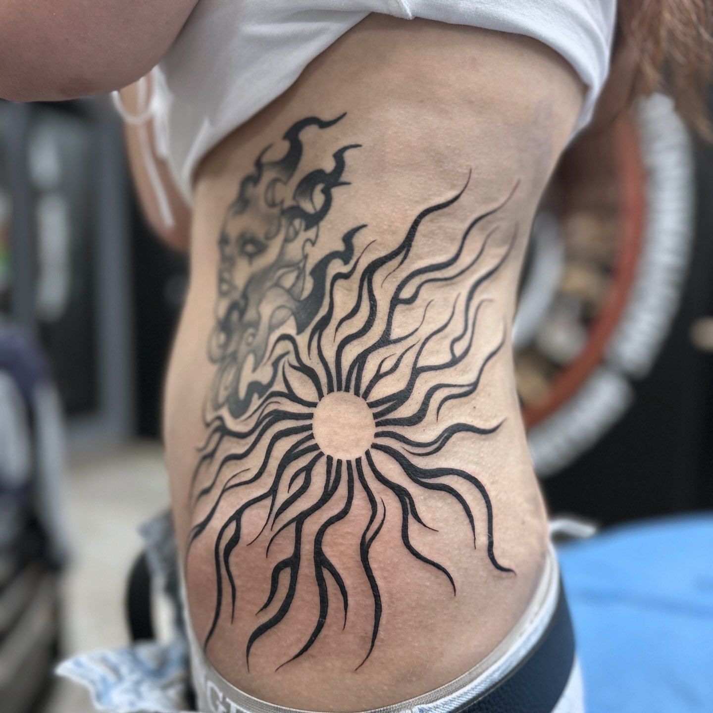 Tattoo on Ribcage Sunburst With Flowing Lines and a Face in Dark Ink.— Whitsunday Tattooing Airlie Beach in Airlie Beach, QLD