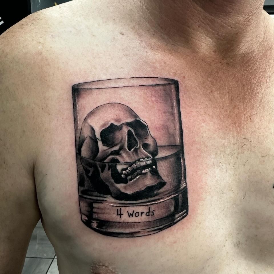 Tattoo of a skull submerged in a glass of liquid, text “4 Words” at base. On a person's chest. — Whitsunday Tattooing Airlie Beach in Whitsundays, QLD