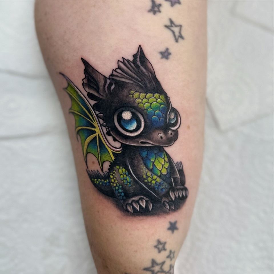 Tattoo of a black, baby dragon with large blue eyes, green-blue scales, and green wings; on an arm, with stars. — Whitsunday Tattooing Airlie Beach in Whitsundays, QLD