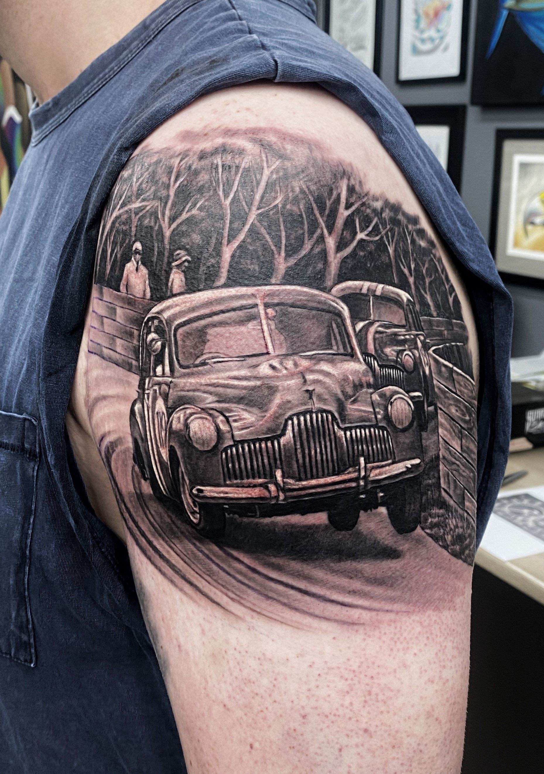 Tattoo of vintage car on arm; black and white scene of car on a road, with trees and figures in the background. — Whitsunday Tattooing Airlie Beach in Whitsundays, QLD