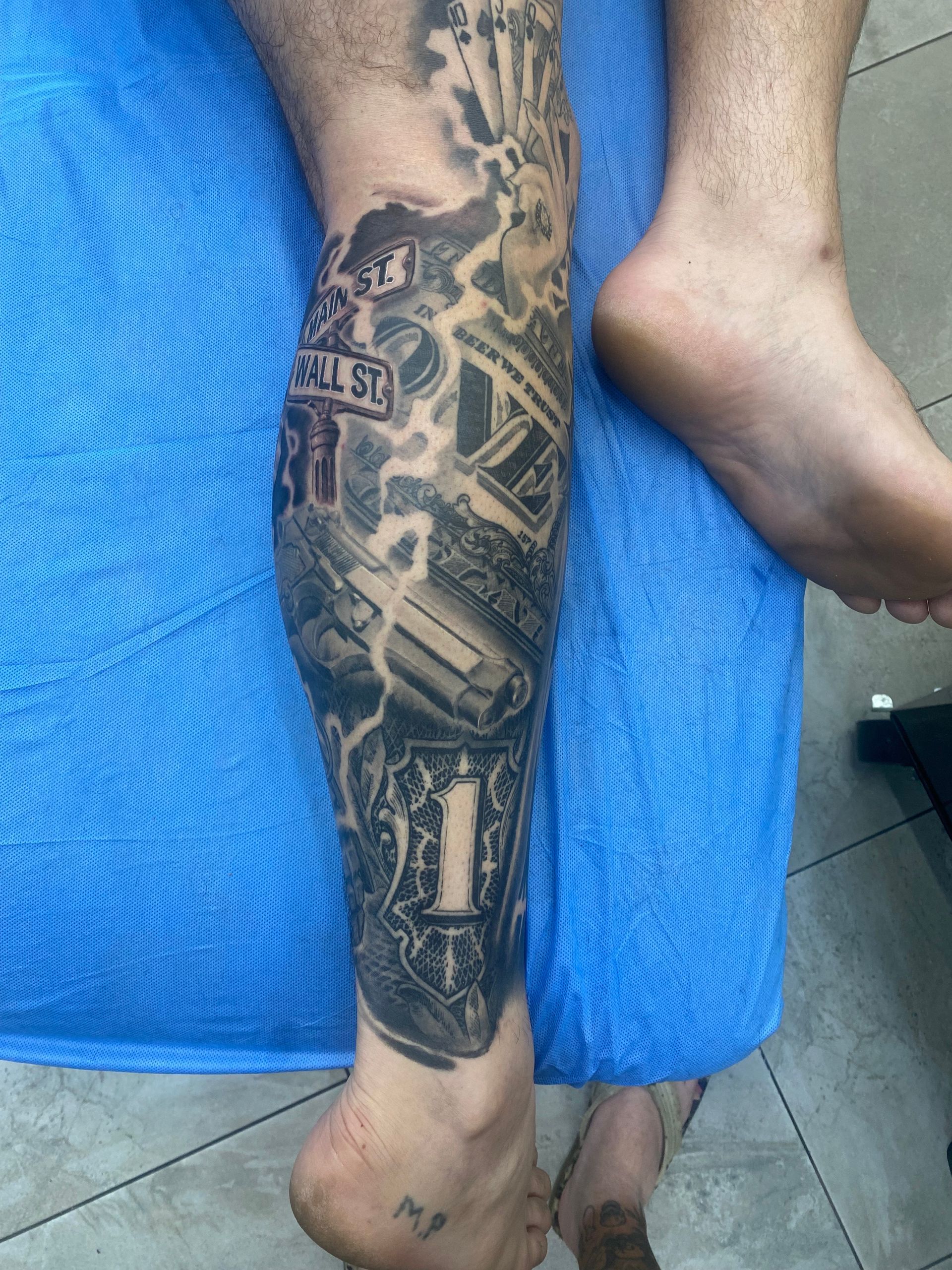 Tattoo on a lower leg featuring a street sign, money, and other elements. Light skin on a blue surface. — Whitsunday Tattooing Airlie Beach in Whitsundays, QLD