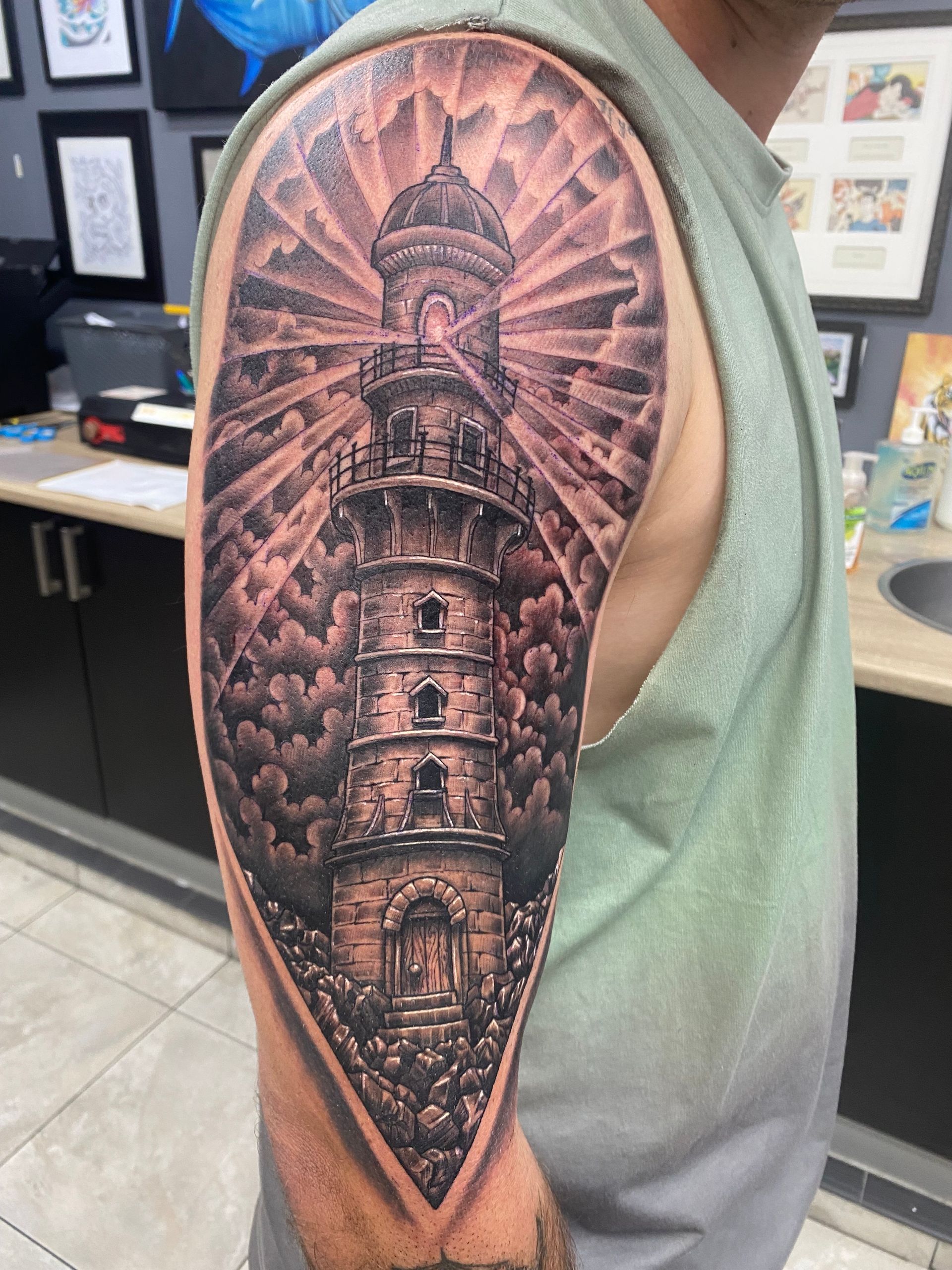 Tattoo of a stone lighthouse with light beams piercing through clouds on a person's upper arm. — Whitsunday Tattooing Airlie Beach in Whitsundays, QLD