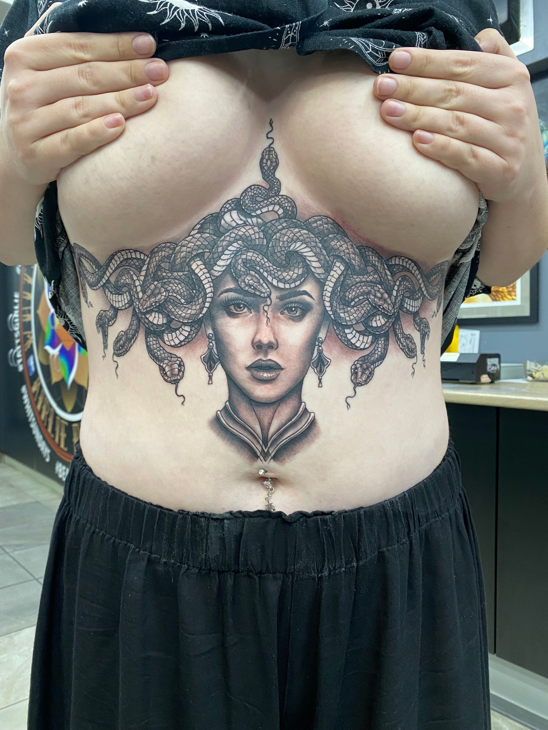 Woman with Medusa chest tattoo, holding up top to reveal the artwork. Tattoo is black and gray, in a studio setting. — Whitsunday Tattooing Airlie Beach in Whitsundays, QLD
