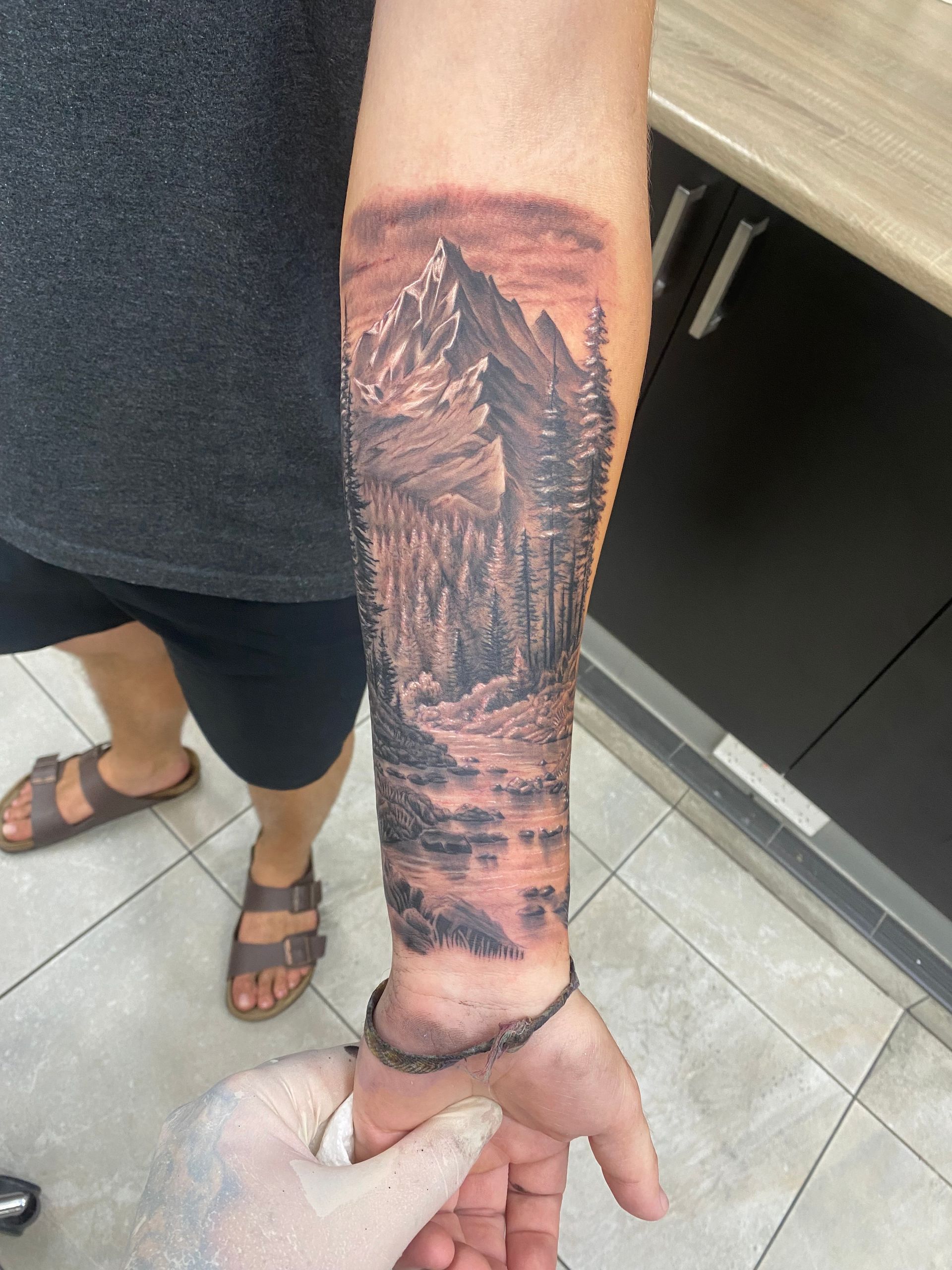 Tattoo of a mountain scene on a forearm. Includes mountain, forest, river, and shaded grayscale ink. — Whitsunday Tattooing Airlie Beach in Whitsundays, QLD