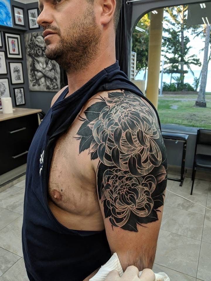 Man with a black floral tattoo on his shoulder and arm, wearing a black tank top. Indoors, sunny outdoor view. — Whitsunday Tattooing Airlie Beach in Whitsundays, QLD
