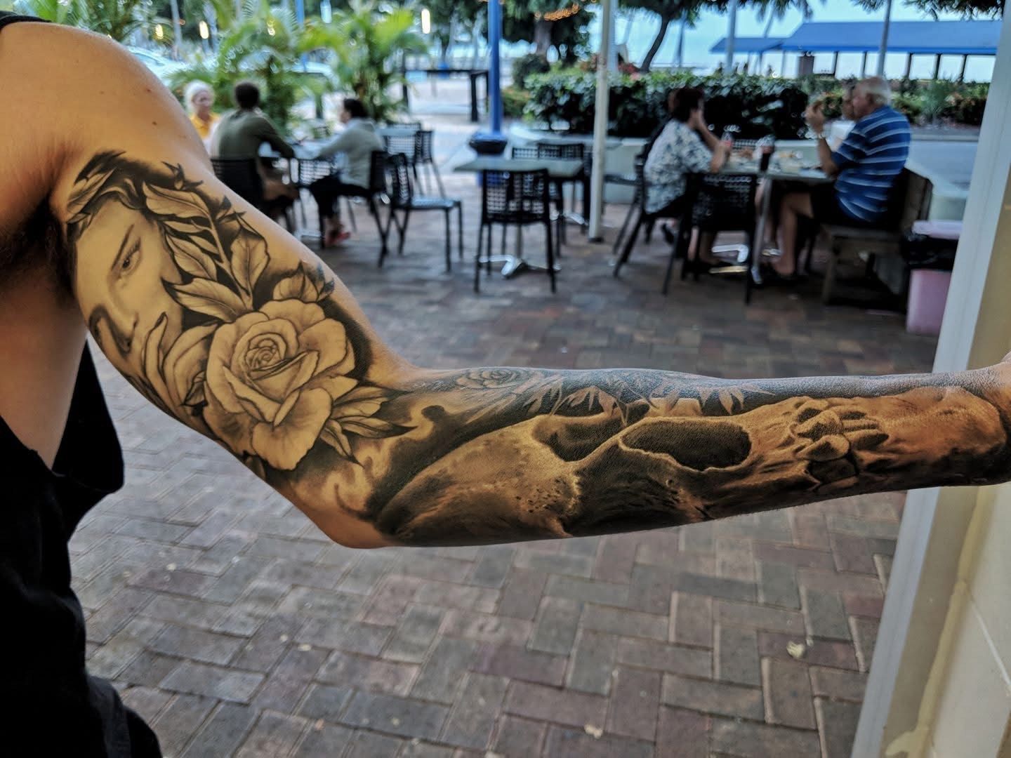 Arm with black and gray floral and skull tattoos, raised in front of a patio dining area. — Whitsunday Tattooing Airlie Beach in Whitsundays, QLD