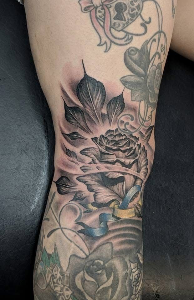 Black and gray rose tattoo on a knee, with surrounding leaves and ribbon details. — Whitsunday Tattooing Airlie Beach in Whitsundays, QLD