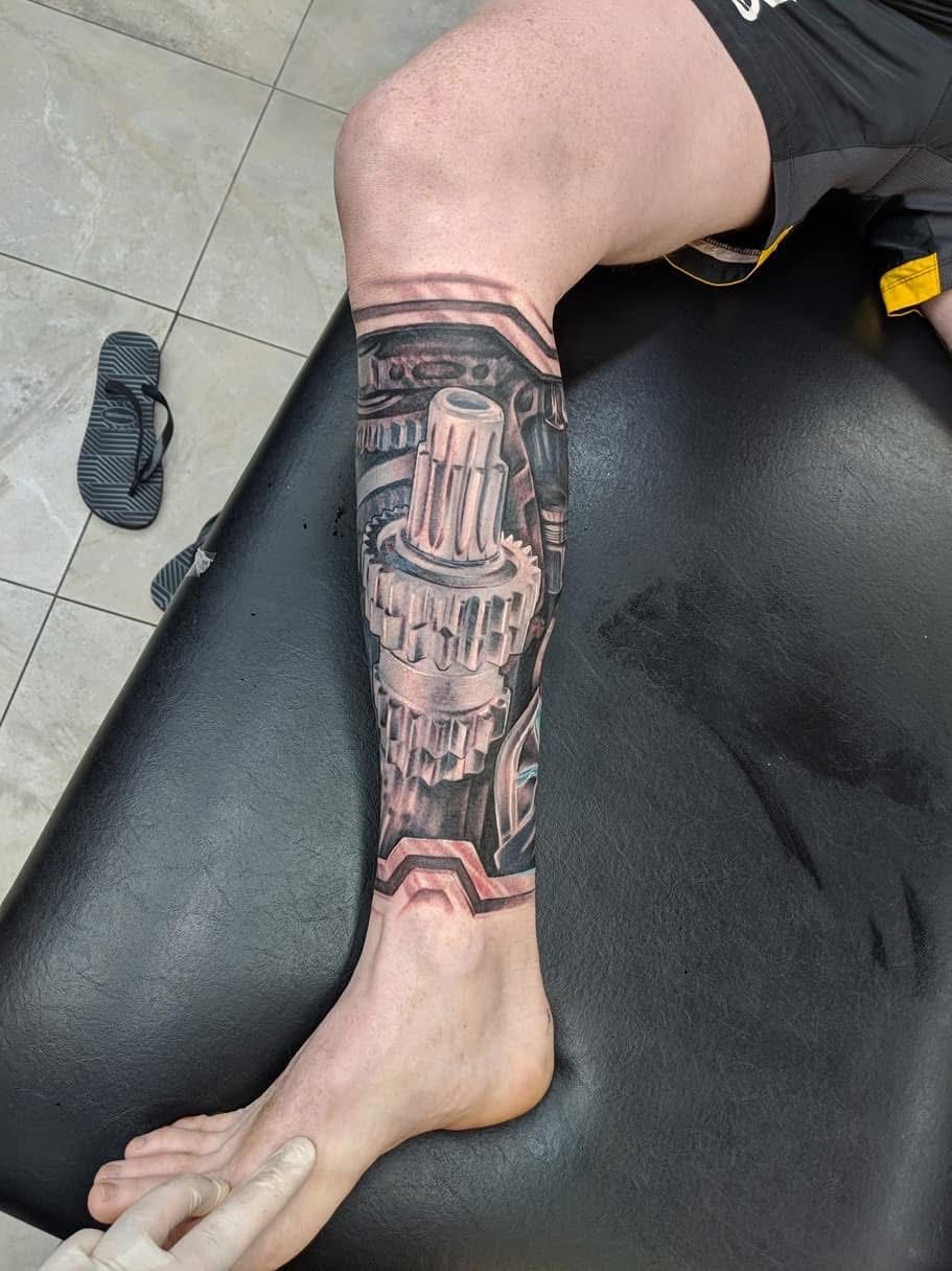 Tattoo of gears and machinery on a person's calf. Leg on black surface, flip flop visible, being worked on. — Whitsunday Tattooing Airlie Beach in Whitsundays, QLD