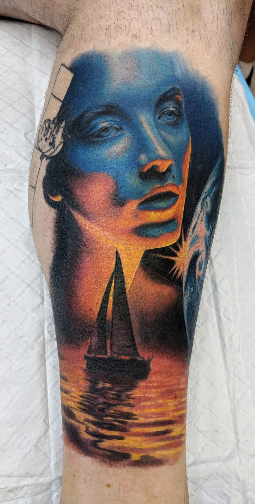 Tattoo on leg: blue face with sunset, sailboat, and planet in a surreal landscape. — Whitsunday Tattooing Airlie Beach in Whitsundays, QLD