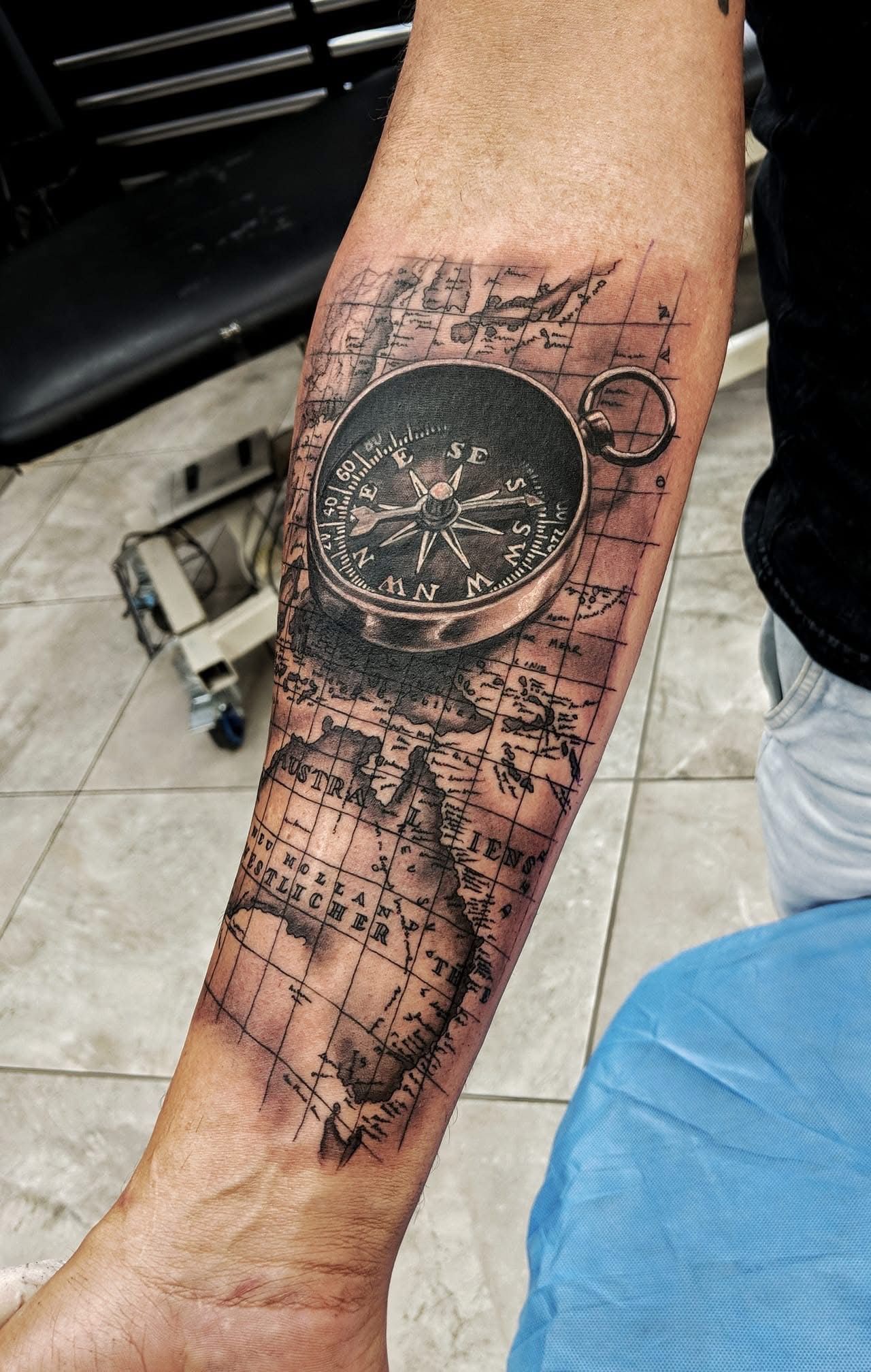 Tattoo of a compass over a map on a person's forearm. Black ink with shading, on a tiled floor. — Whitsunday Tattooing Airlie Beach in Whitsundays, QLD
