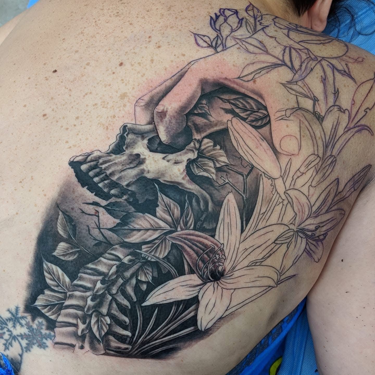 Tattoo on back: Skull in leaves, hand holding it, outlined flowers extending from shoulder. — Whitsunday Tattooing Airlie Beach in Whitsundays, QLD
