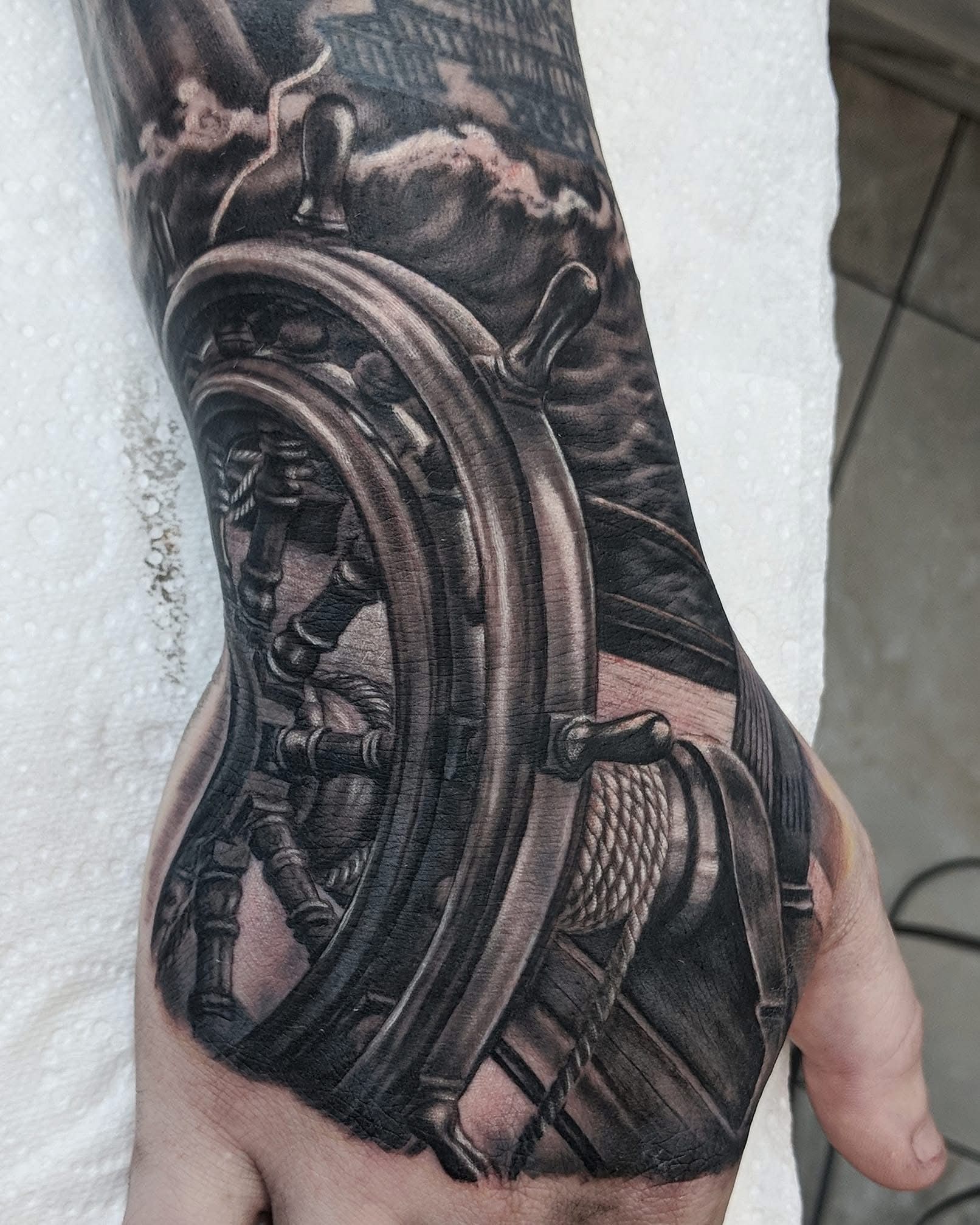 Black and grey tattoo of a ship's wheel on a hand, with a stormy sea and ship in the background. — Whitsunday Tattooing Airlie Beach in Whitsundays, QLD