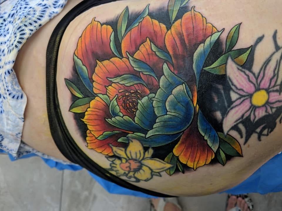 Colorful peony tattoo on a person's hip; orange, blue, and green petals, with a smaller flower nearby. — Whitsunday Tattooing Airlie Beach in Whitsundays, QLD