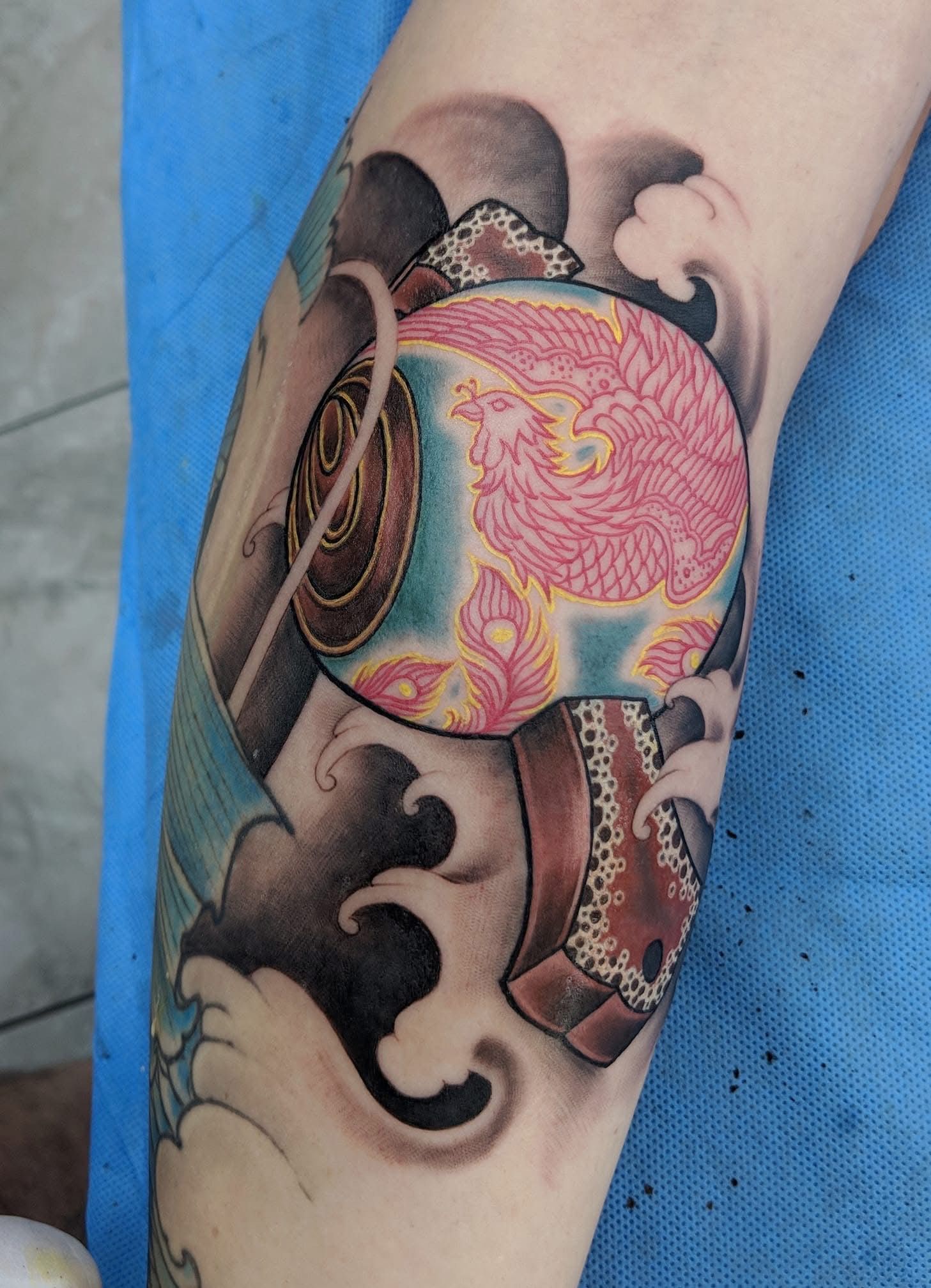Tattoo on arm depicting a pink phoenix on a turquoise and brown drum with water waves and blue accents.