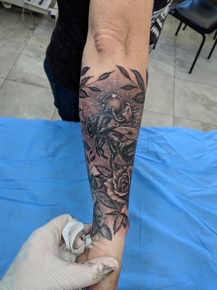Arm with a grayscale tattoo of a bee, roses, and leaves. A gloved hand touches the arm. Blue cloth backdrop. — Whitsunday Tattooing Airlie Beach in Whitsundays, QLD