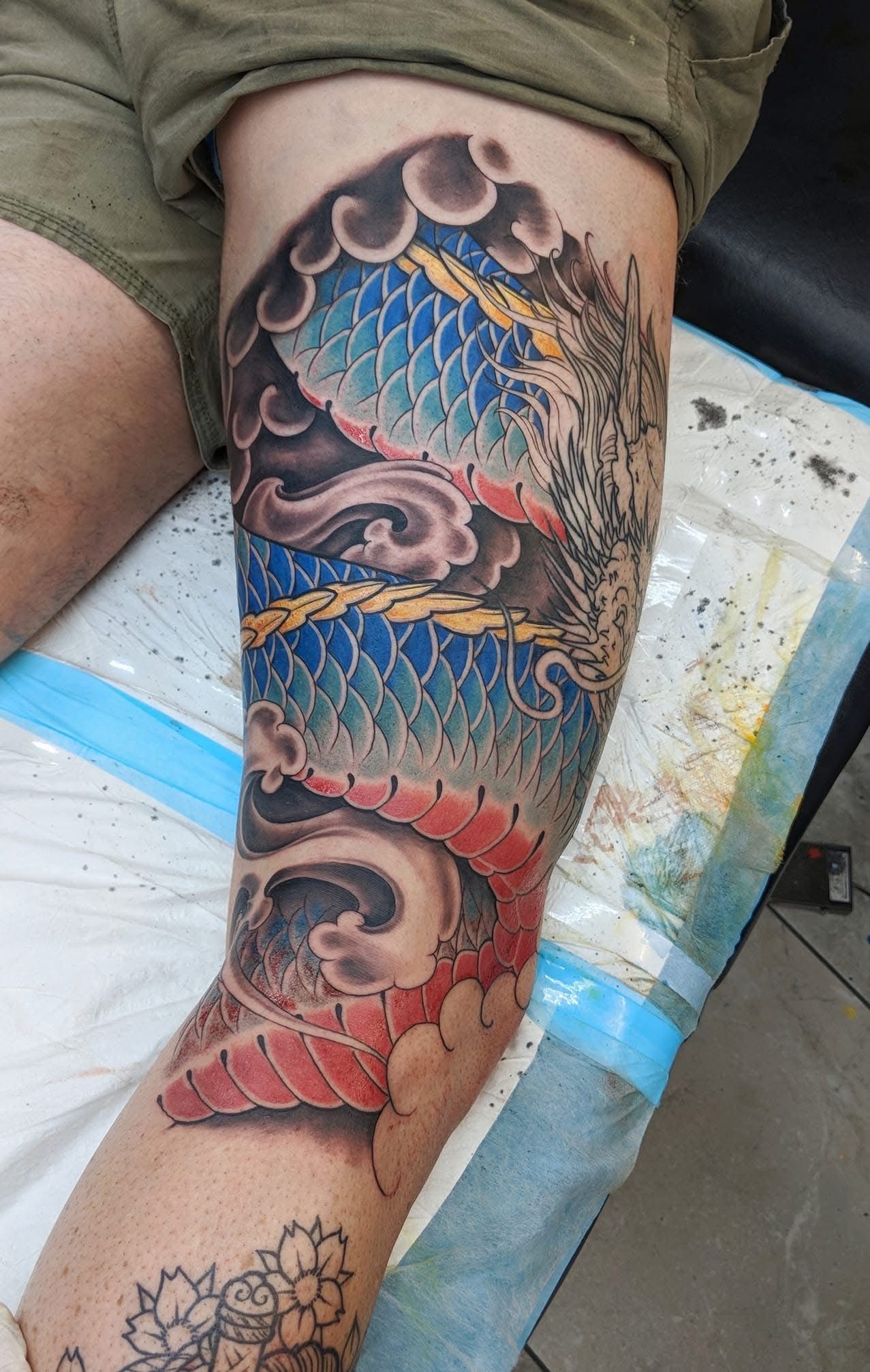 Tattoo of a dragon on a leg. Blue, red, and gray scales, swirling clouds, and waves. — Whitsunday Tattooing Airlie Beach in Whitsundays, QLD