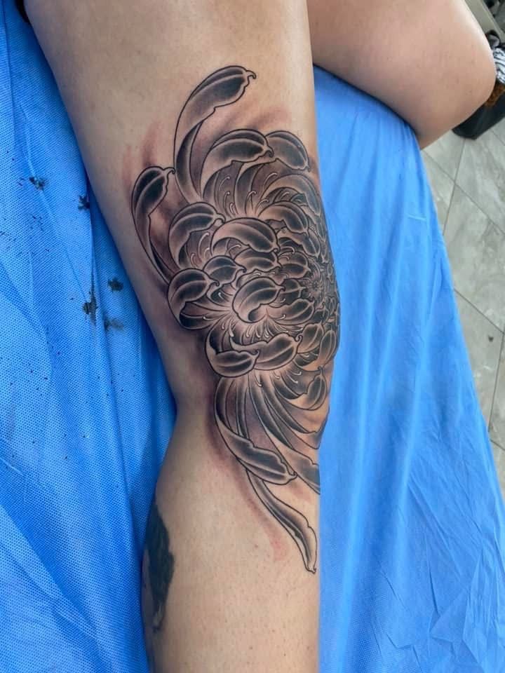 Black and gray chrysanthemum tattoo on a knee, partially shaded, lying on a blue surface. — Whitsunday Tattooing Airlie Beach in Whitsundays, QLD