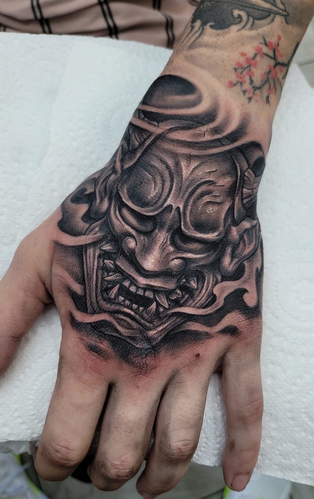 Detailed hand tattoo of a demon mask, shaded in black and gray, with surrounding smoky effects. — Whitsunday Tattooing Airlie Beach in Whitsundays, QLD