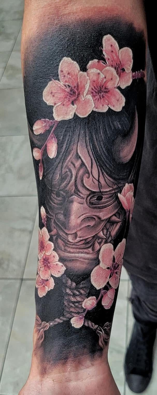 Tattoo of a Japanese demon mask with cherry blossoms on an arm. Dark background. — Whitsunday Tattooing Airlie Beach in Whitsundays, QLD