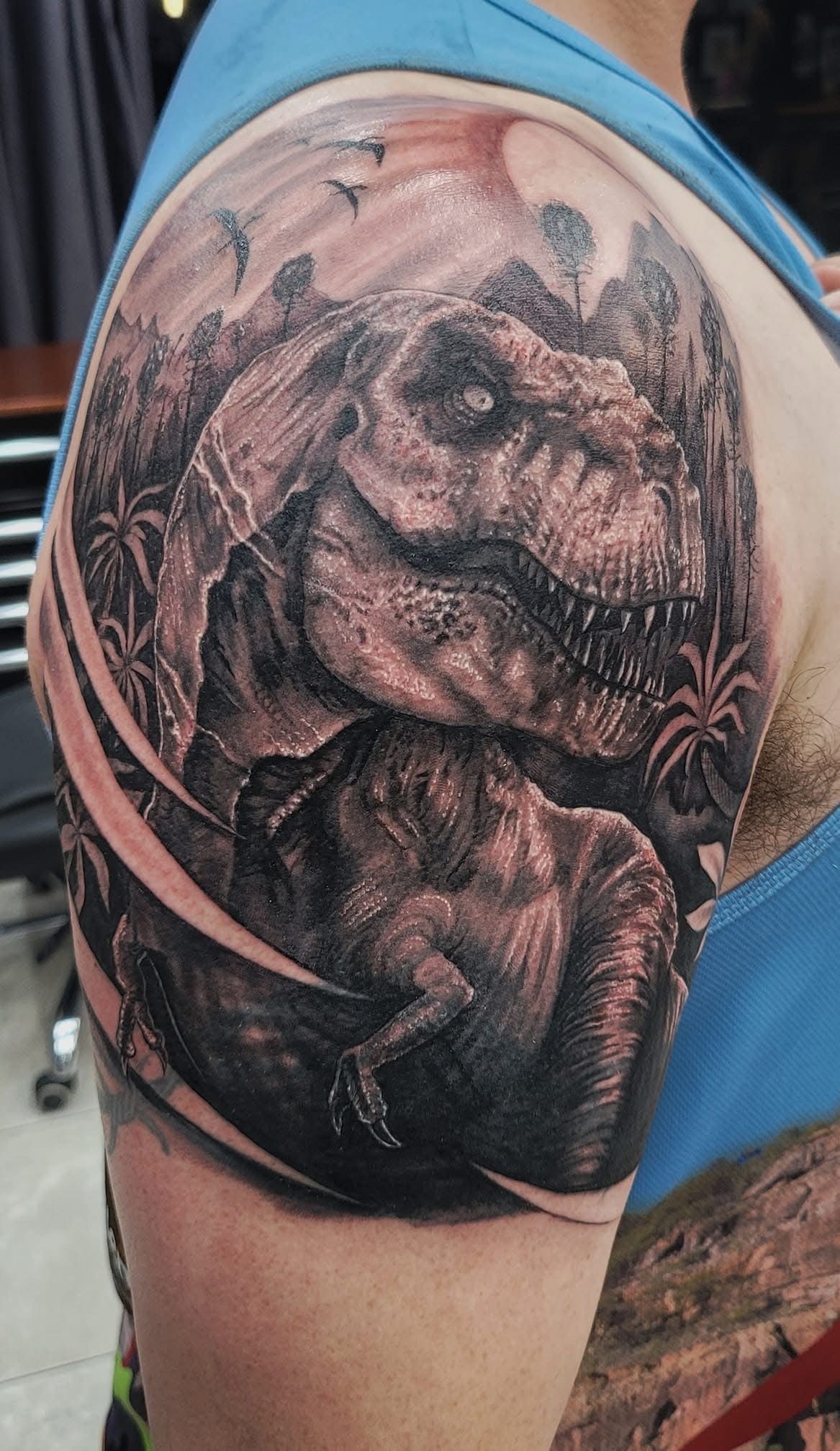 Tattoo of a detailed T-Rex on a shoulder, dark grey shading, with a lush jungle background. — Whitsunday Tattooing Airlie Beach in Whitsundays, QLD