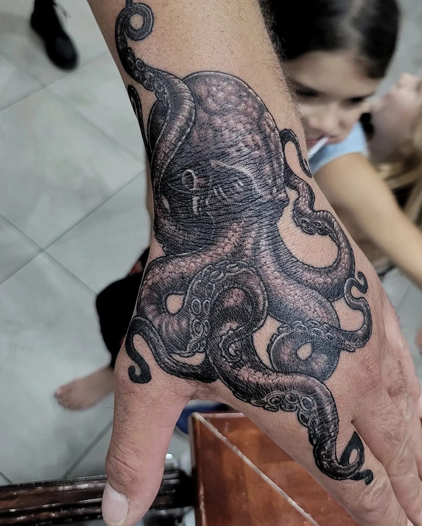 Hand tattoo of a dark octopus on a person's palm, with blurred children in the background. — Whitsunday Tattooing Airlie Beach in Whitsundays, QLD