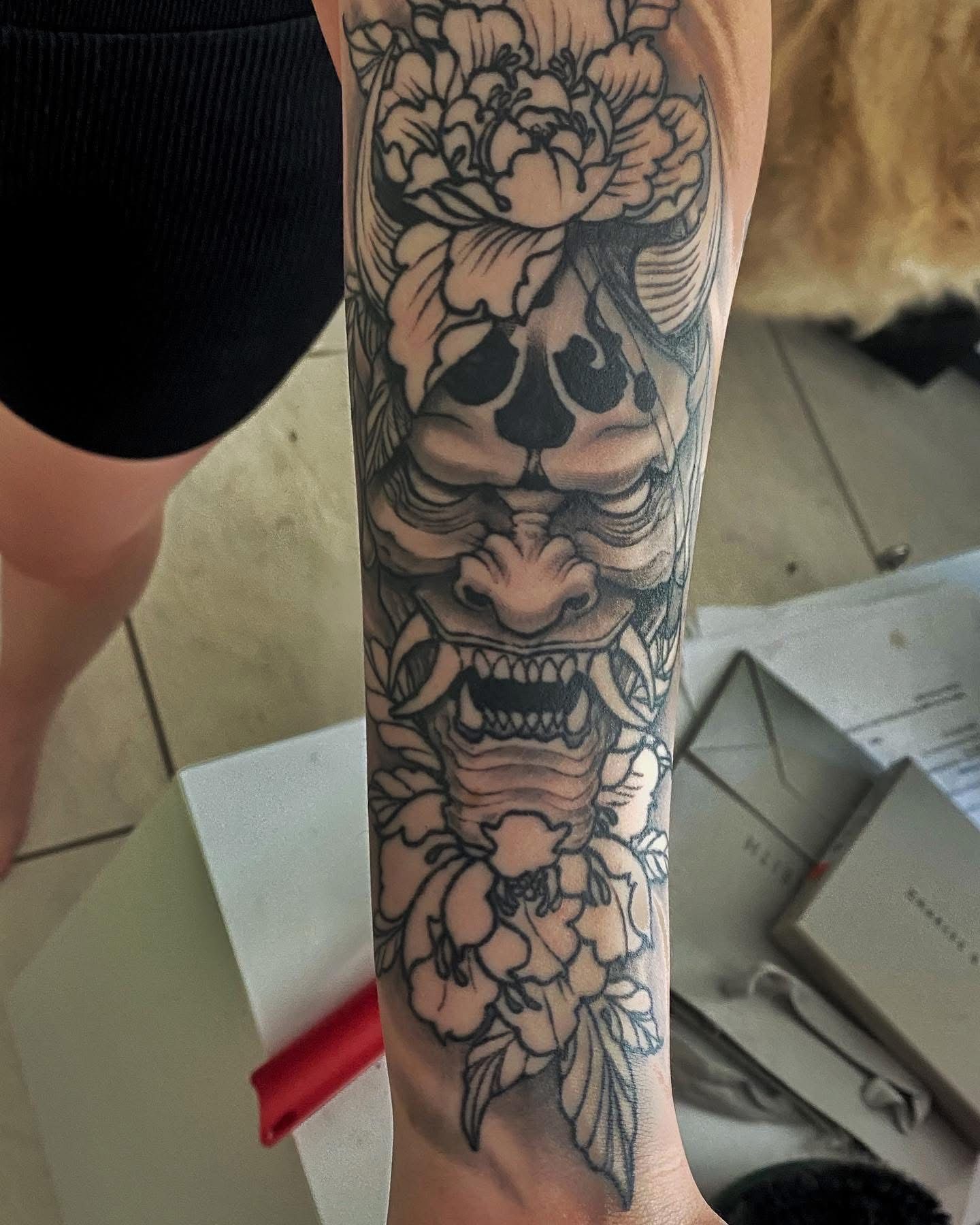 Tattoo of a Japanese demon mask with peony flowers on a forearm. Black ink, gray shading. — Whitsunday Tattooing Airlie Beach in Whitsundays, QLD